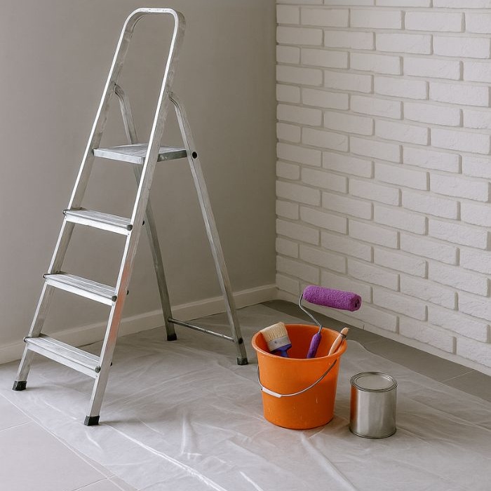 Escabeau métallique, seau orange et rouleaux de peinture violets posés sur bâche plastique dans une pièce en rénovation avec mur en briques blanches.