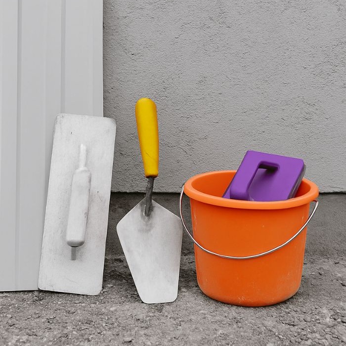 Outils de ravalement de façade posés sur le sol : taloche, truelle à manche jaune et seau orange avec platoir violet, sur fond de mur gris.